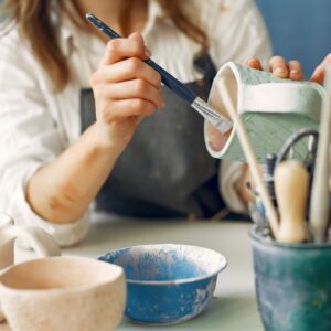 Crop anonymous craftswoman in apron painting handmade ceramic products while working in contemporary creative pottery
