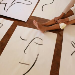 From above of faceless barefoot casually dressed woman with brush sitting on floor while placed artworks in abstract style